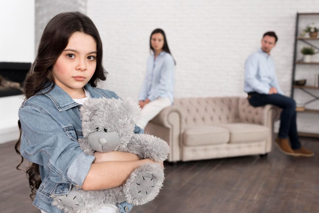 Menina triste segurando ursinho com pais brigados ao fundo
