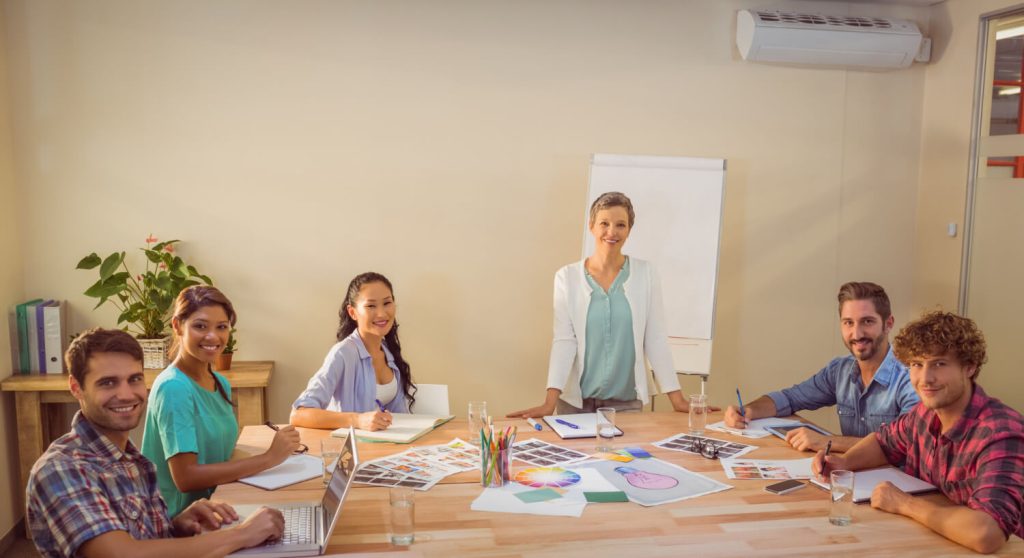 Mulher de mais idade ao meio de uma mesa, em reunião com profissionais de educação mais jovens.